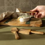 Person using a wooden cheese knife to cut into a wheel of cheese on a wooden board.