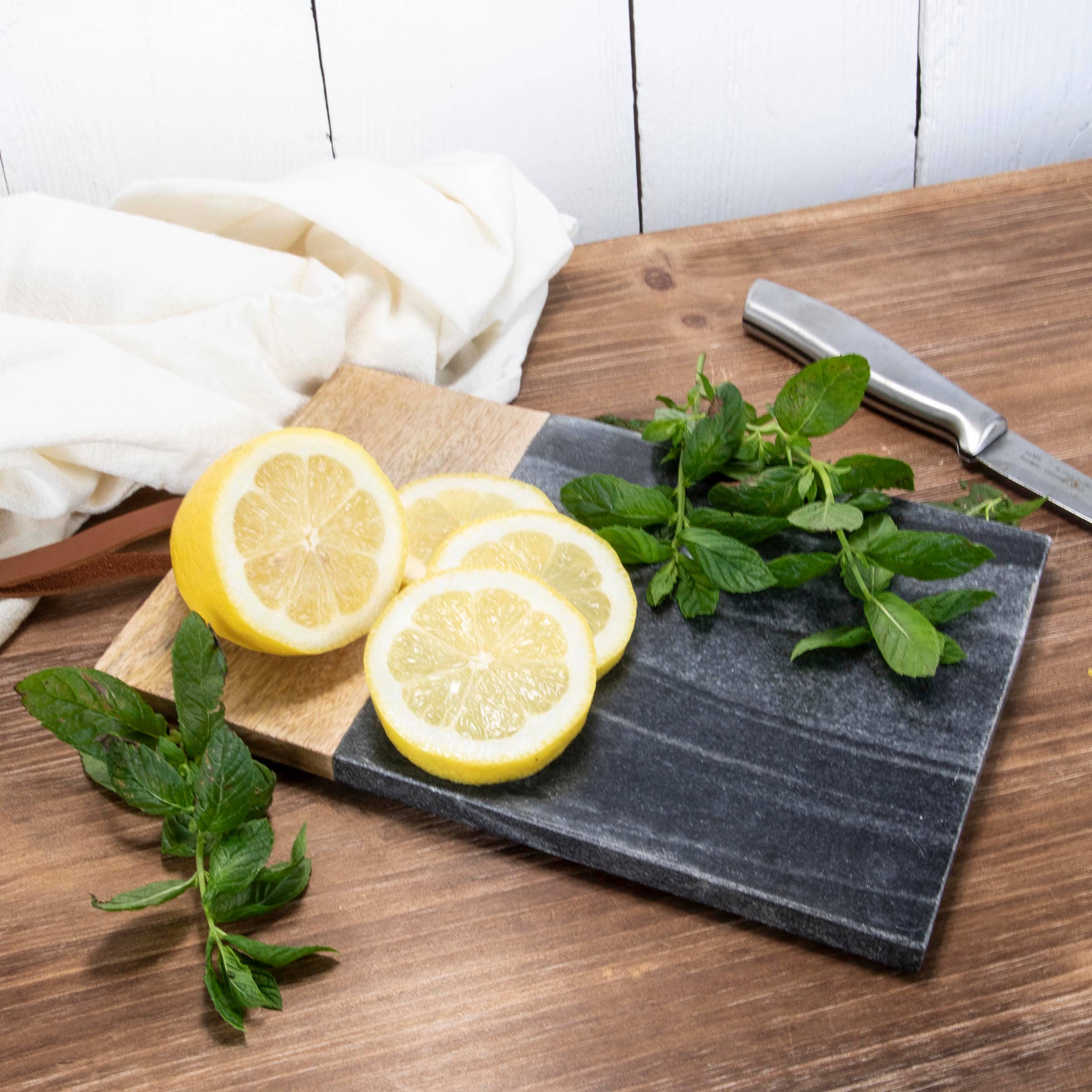 Sliced lemons and fresh mint leaves on a dark marble cutting board with a knife, on a wooden surface.
