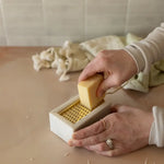 Person using a soap dish with cheese grater and block of cheese on a wooden surface.