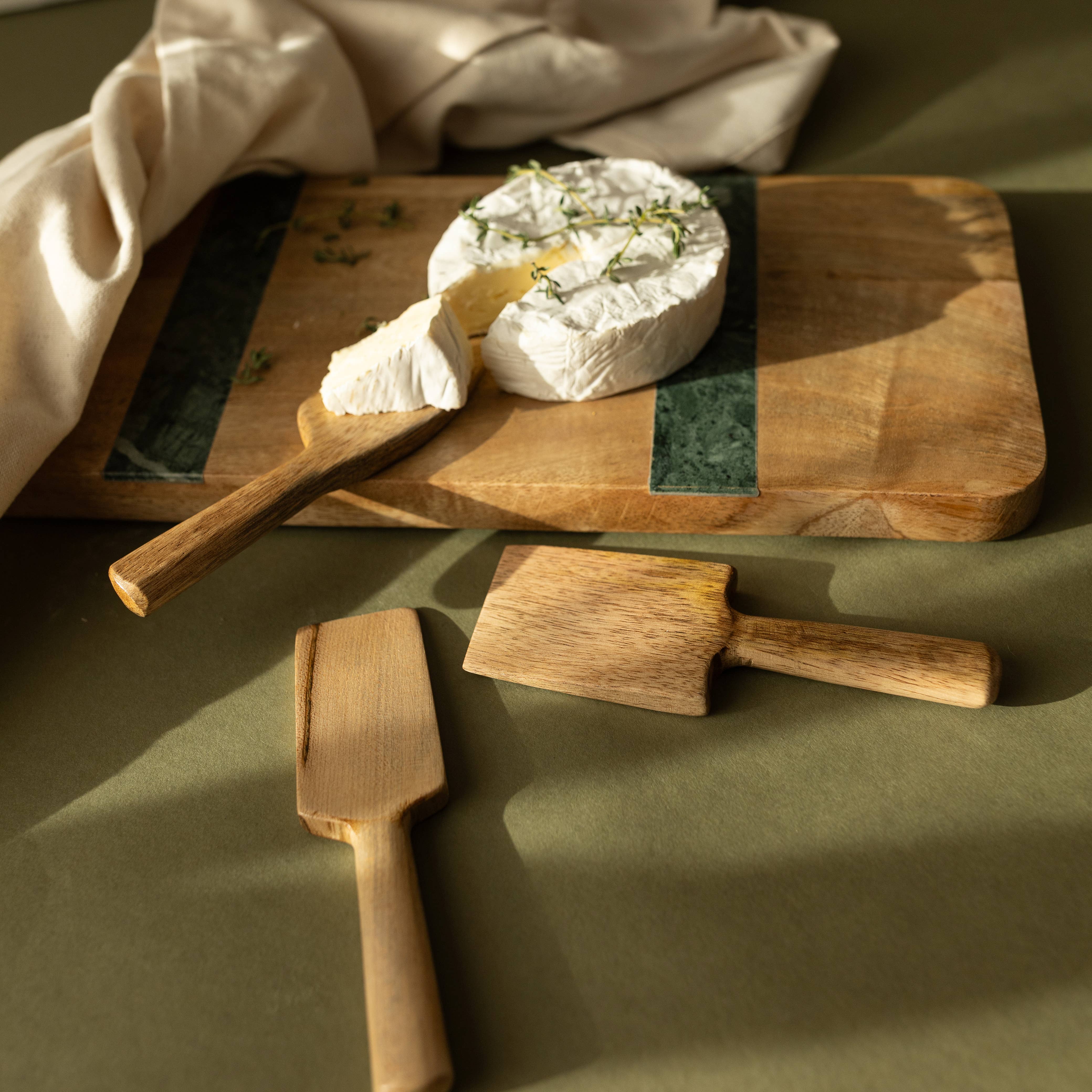 Wooden cutting board with a wheel of cheese and two wooden cheese knives on a green surface.