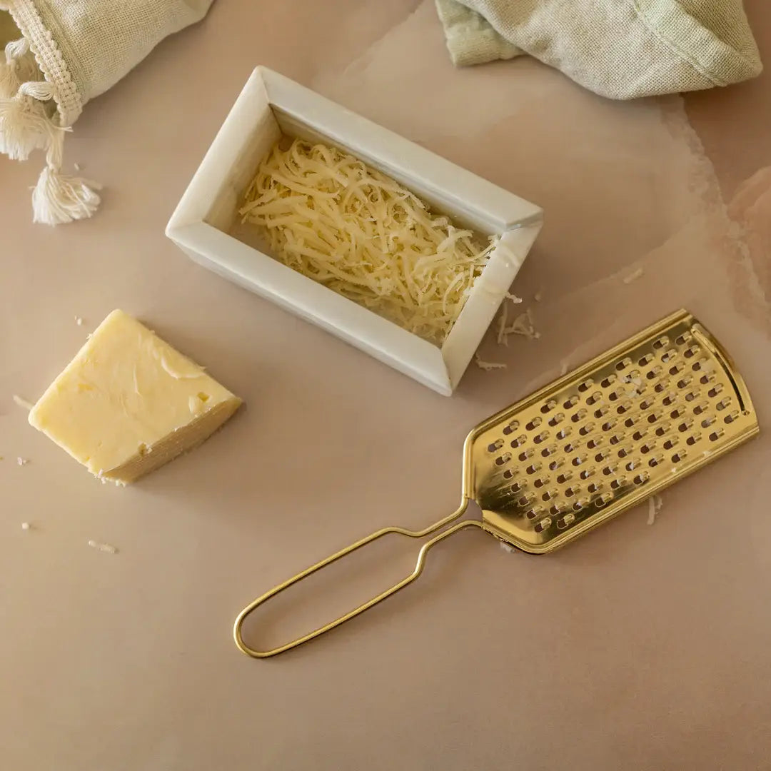 Gold grater next to a block of cheese and a container of stringy cheese on a beige surface.