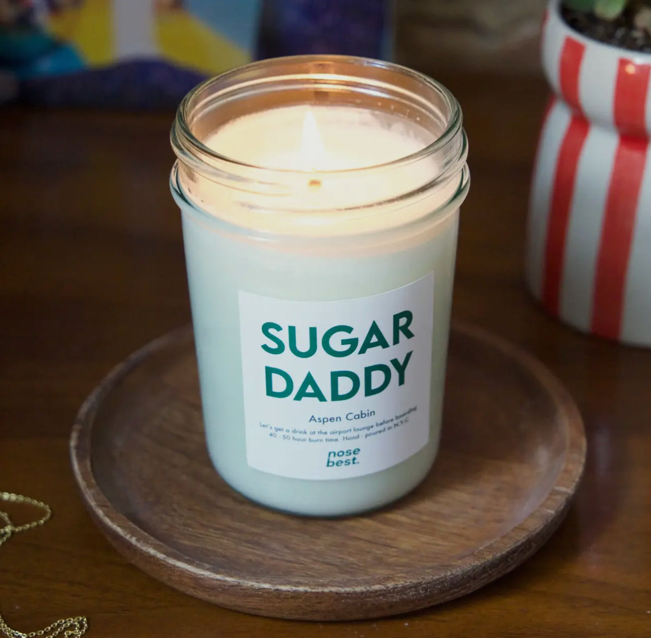 Candle in a jar labeled 'SUGAR DADDY' on a wooden coaster with a colorful background.