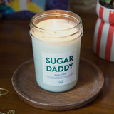 Candle in a jar labeled 'SUGAR DADDY' on a wooden coaster with a colorful background.