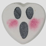 Heart-shaped bath bomb with black oval eyes and mouth with pink dots for blush on a light gray background