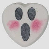 Heart-shaped bath bomb with black oval eyes and mouth with pink dots for blush on a light gray background