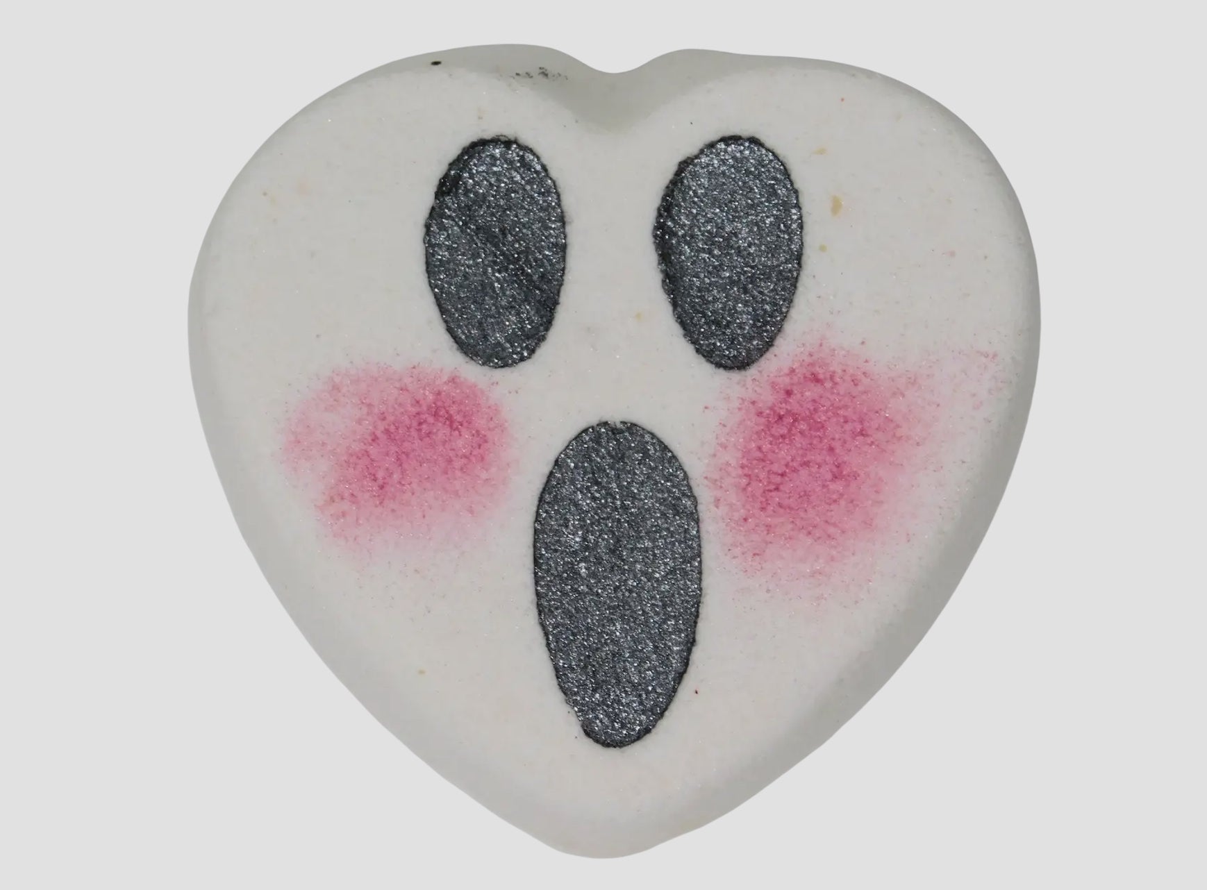 Heart-shaped bath bomb with black oval eyes and mouth with pink dots for blush on a light gray background