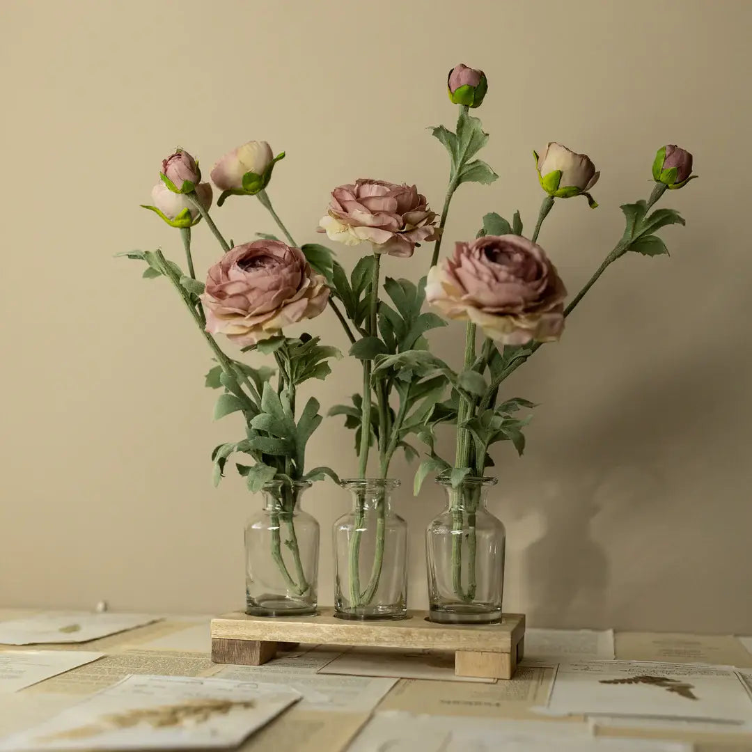 Three glass vases with pink flowers on a wooden stand against a beige wall.