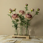 Three glass vases with pink flowers on a wooden stand against a beige wall.