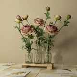 Three glass vases with pink flowers on a wooden stand against a beige wall.