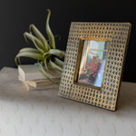 Brass photo frame with a textured pattern on a surface with a plant and books in the background.